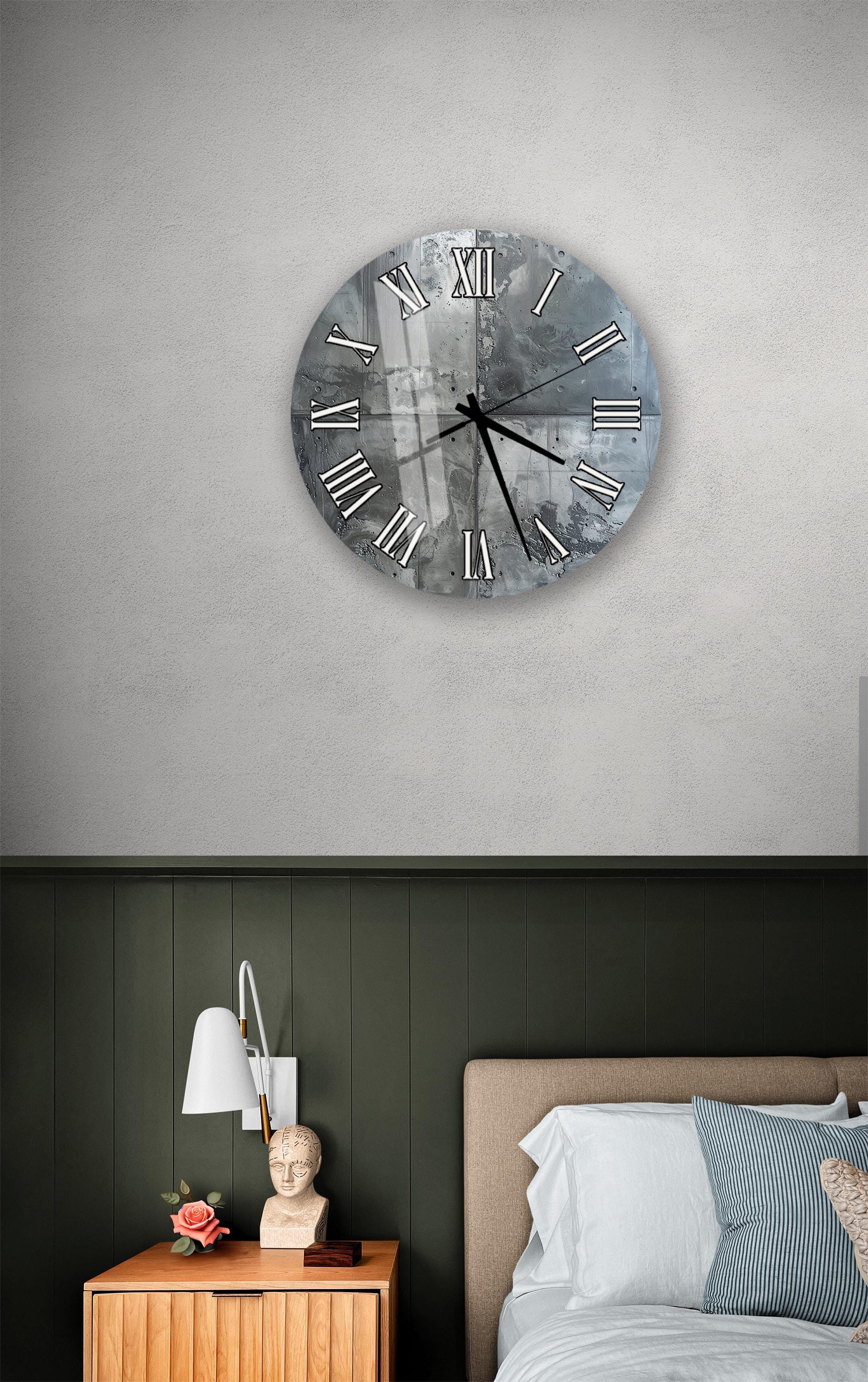 a bedroom setting with a large wall clock hanging above a bed. The clock has a silver and black color scheme and displays Roman numerals. The room has a green wall, a wooden nightstand with a lamp, and a bed with white sheets.