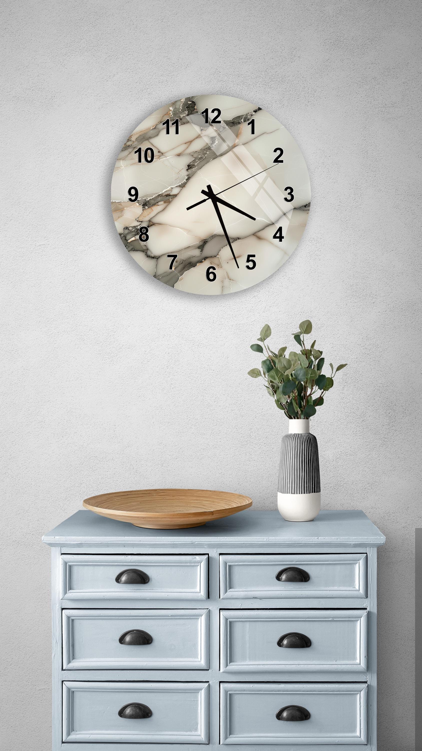 a wall clock with a marble-like design, a wooden bowl, and a vase with greenery on a light blue dresser.