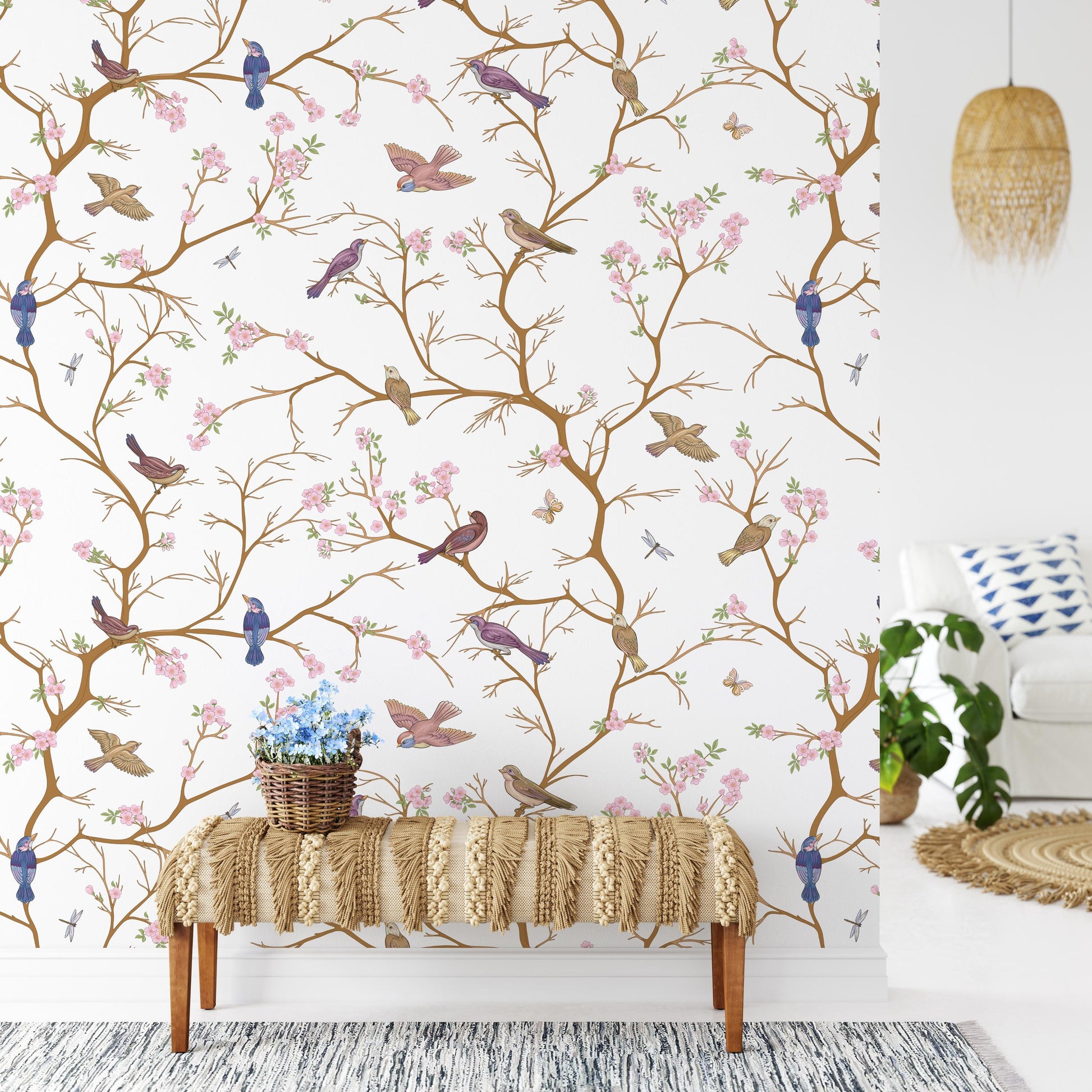 a wallpapered wall with a decorative pattern of birds and flowers, including blue birds and pink flowers. In front of the wall, there is a bench with a woven seat and a basket of flowers on it. The room also includes a white couch, a woven rug, and a potted plant.