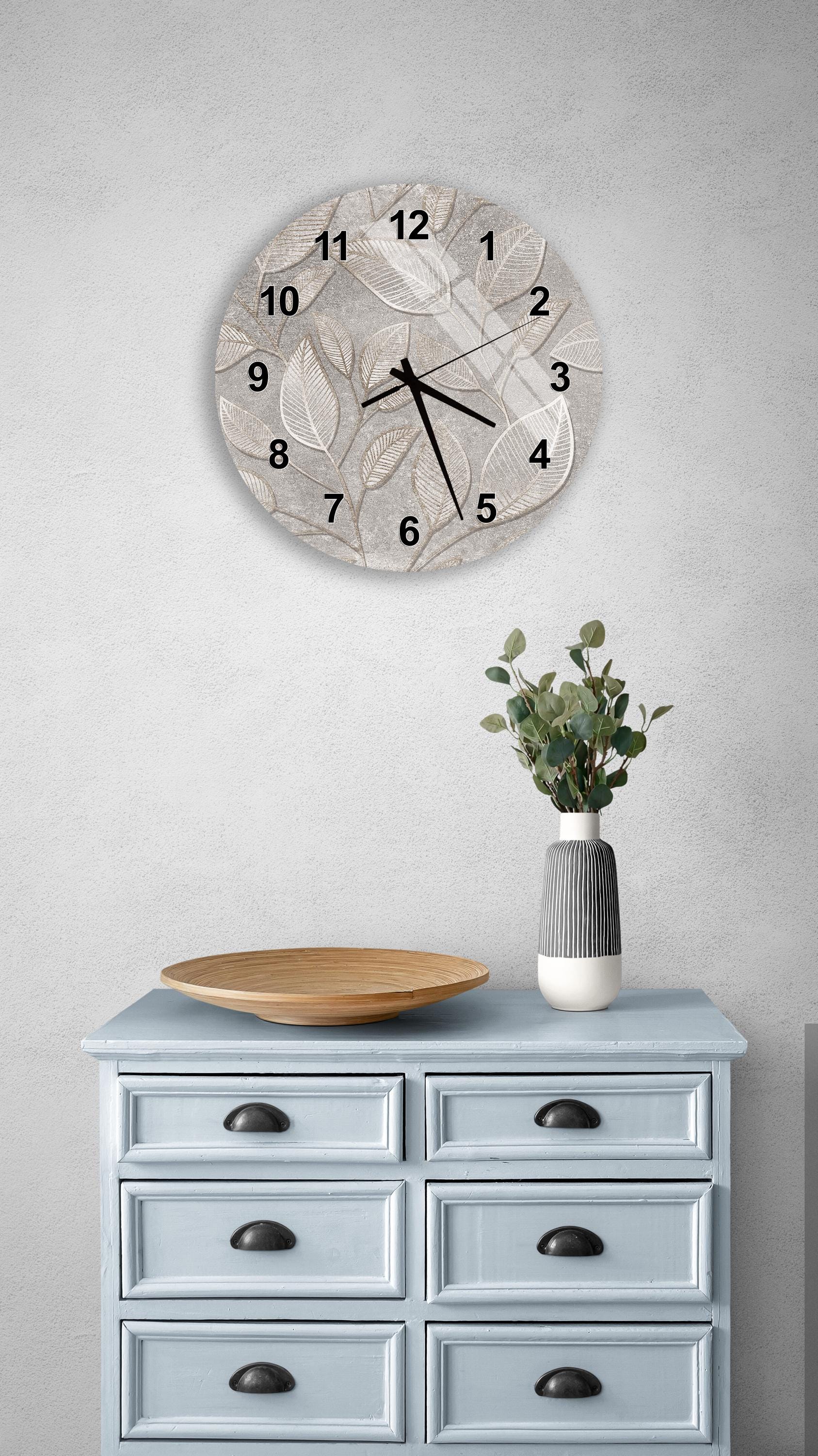 a wall clock with a leaf design, a wooden bowl on a light blue dresser, and a vase with greenery on a gray wall.