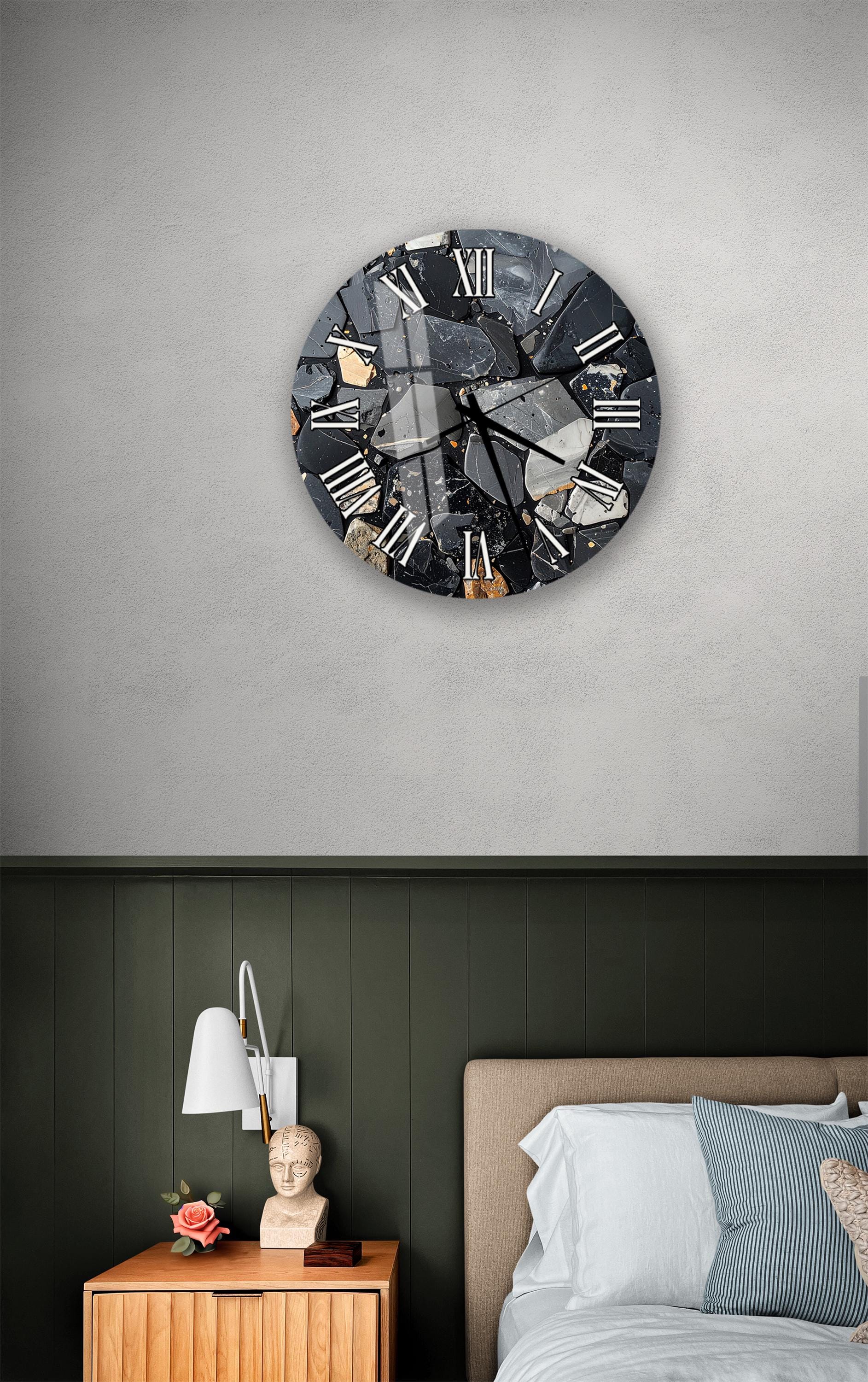 a bedroom setting with a large, round wall clock displaying Roman numerals. The clock is mounted on a wall above a bed with a wooden headboard. The room has a green wall and a wooden nightstand with a lamp and a vase of flowers.
