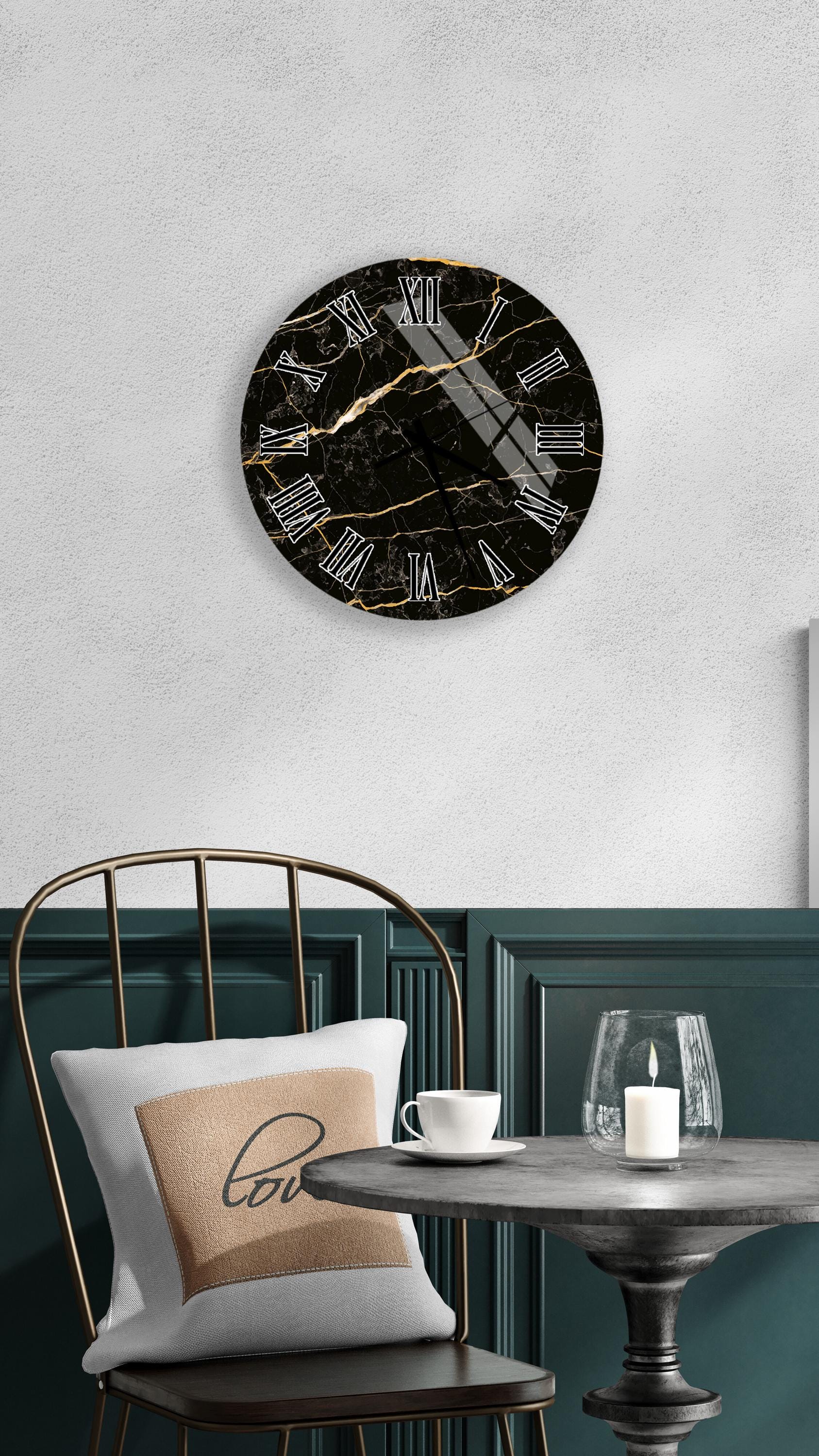a black marble clock with a gold-colored design, mounted on a white wall above a black metal chair. The chair has a white pillow with the word "love" printed on it. In front of the chair, there is a small table with a glass candle holder and a white teacup.