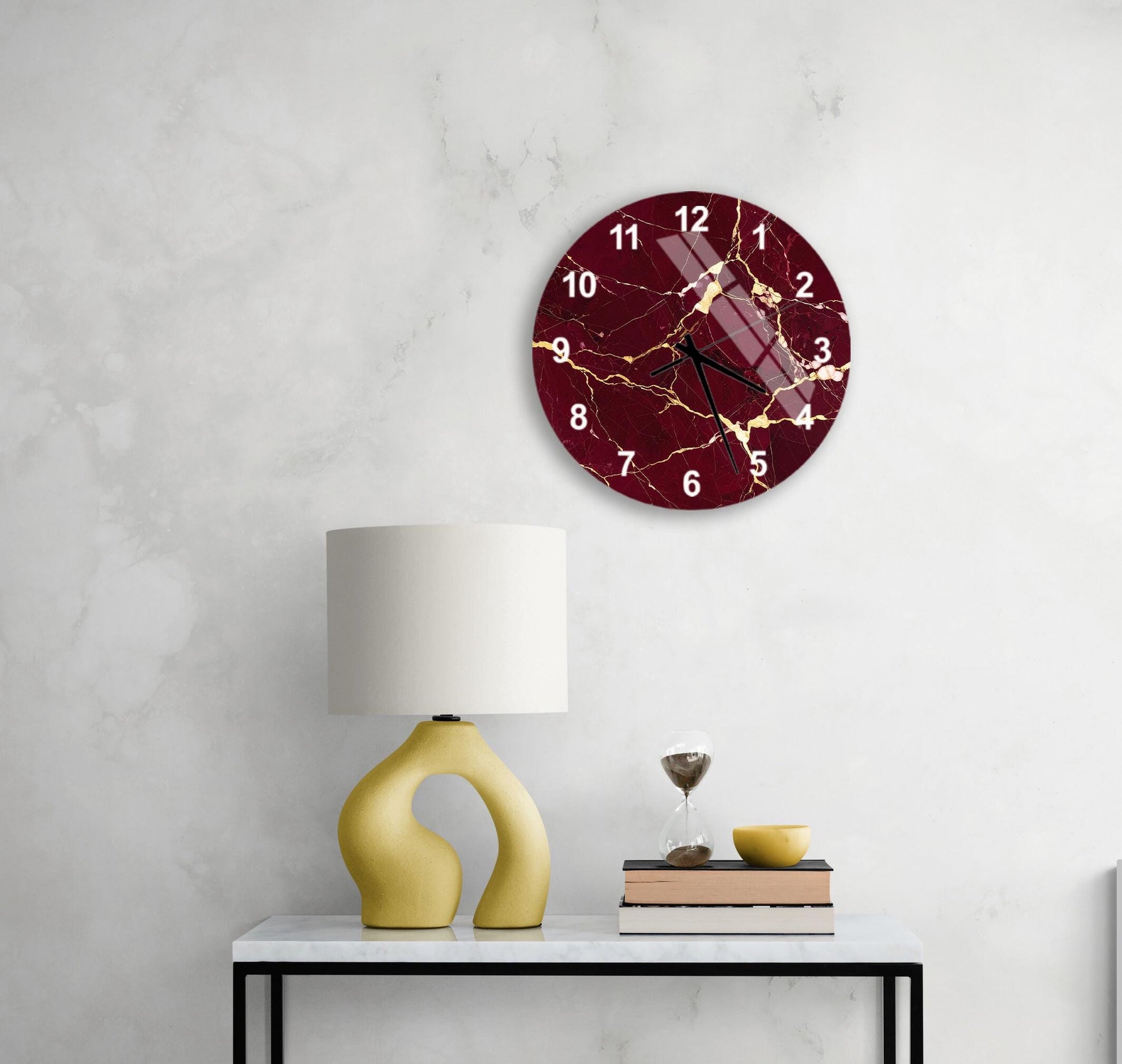 a white marble wall with a round clock featuring a maroon and gold design, a yellow lamp with a unique curved base, a stack of books, and a glass of wine on a white table.