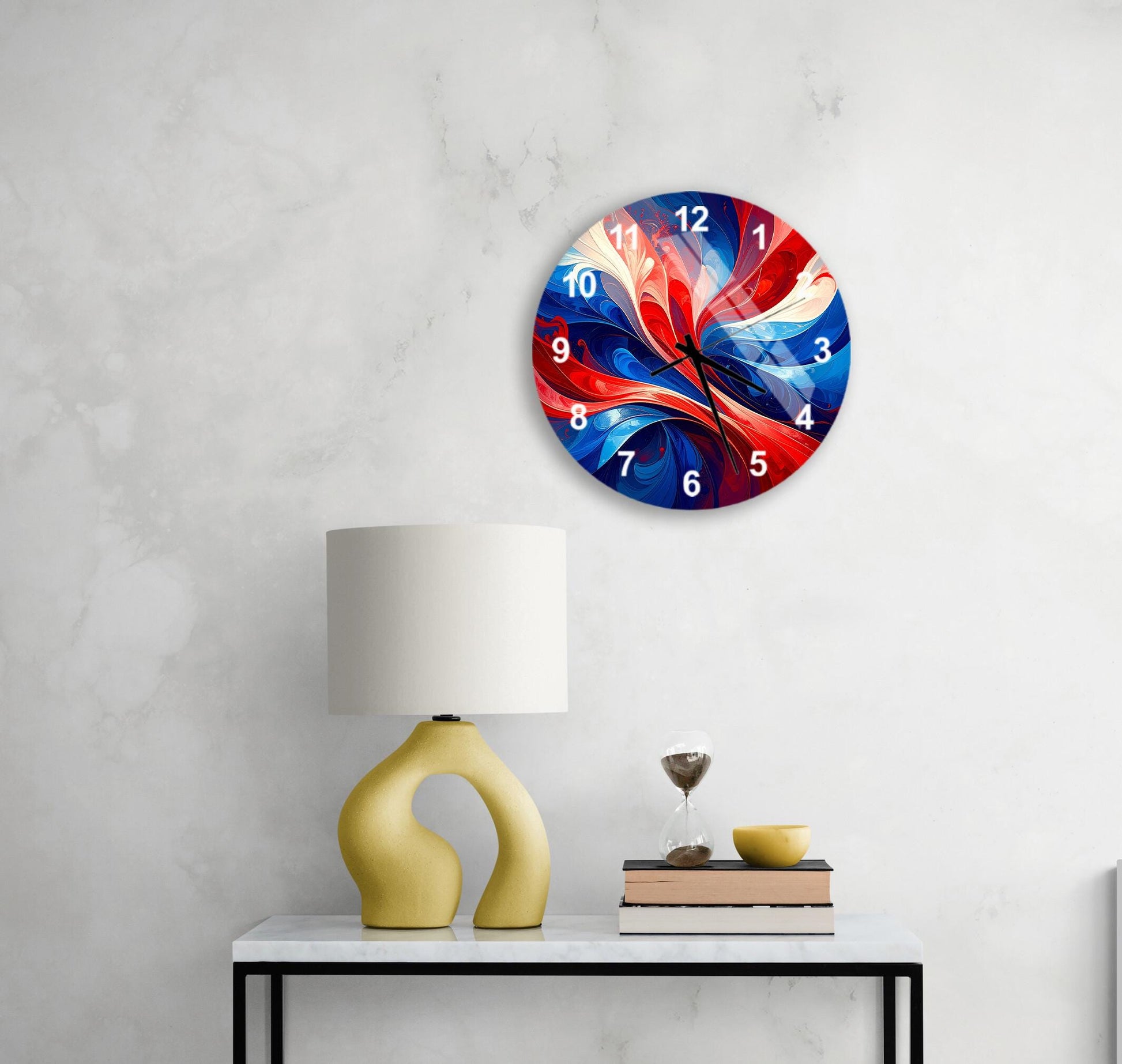 a modern wall clock with a vibrant, abstract design in red, blue, and white colors, hanging above a white table with a yellow lamp and a stack of books.
