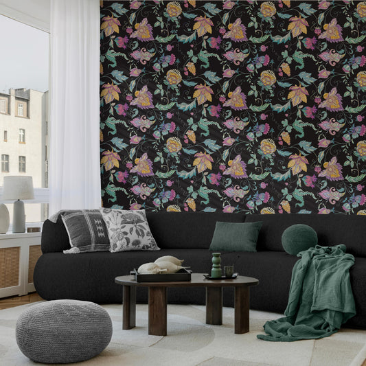 a cozy living room with a black sofa, a wooden coffee table, and a gray ottoman. The wall behind the sofa is covered in a vibrant floral wallpaper, adding a touch of nature to the space. The room is illuminated by natural light coming through a window, creating a warm and inviting atmosphere.
