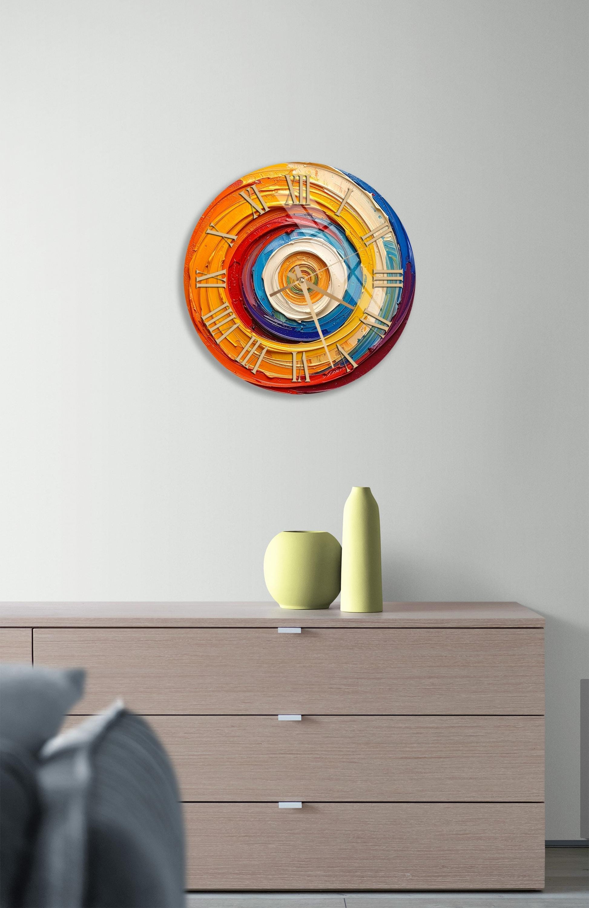 a modern art wall clock with a colorful, abstract design, mounted on a wall above a wooden dresser with a vase and a small sculpture.