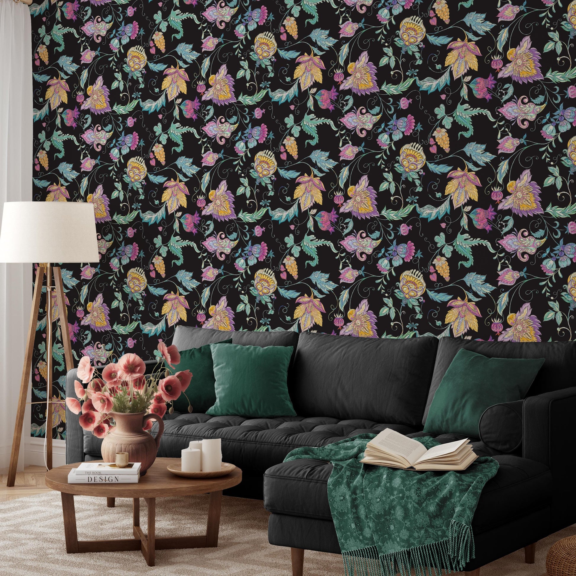 a cozy living room with a black sofa adorned with green throw pillows, a wooden coffee table, and a lamp. The wall behind the sofa is covered in a vibrant floral wallpaper, adding a touch of nature and color to the space. A vase of pink flowers is placed on the coffee table, and an open book is resting on the sofa.
