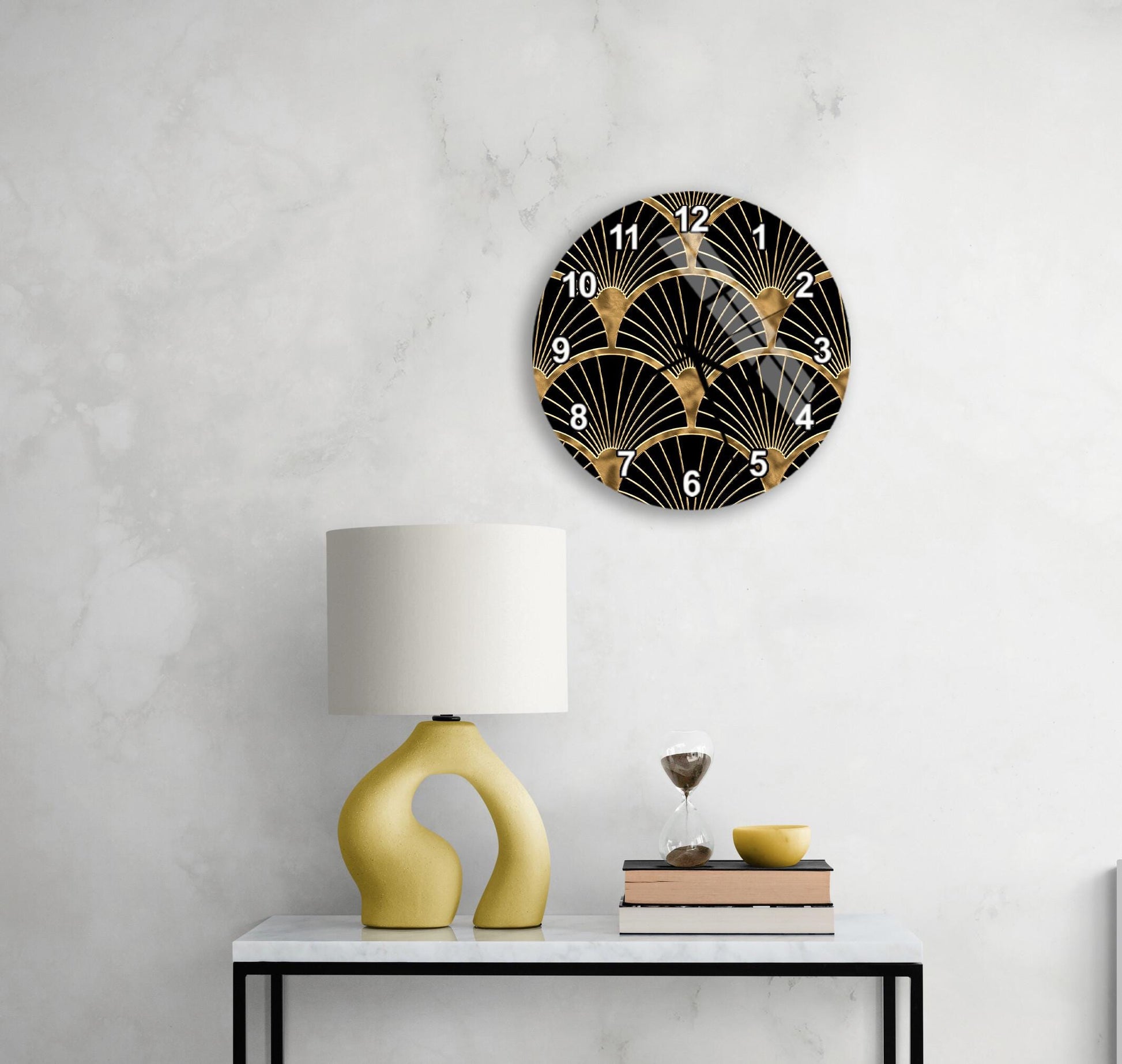 a modern black and gold wall clock with a black and gold design, mounted on a white wall above a white table with a yellow lamp and a stack of books.