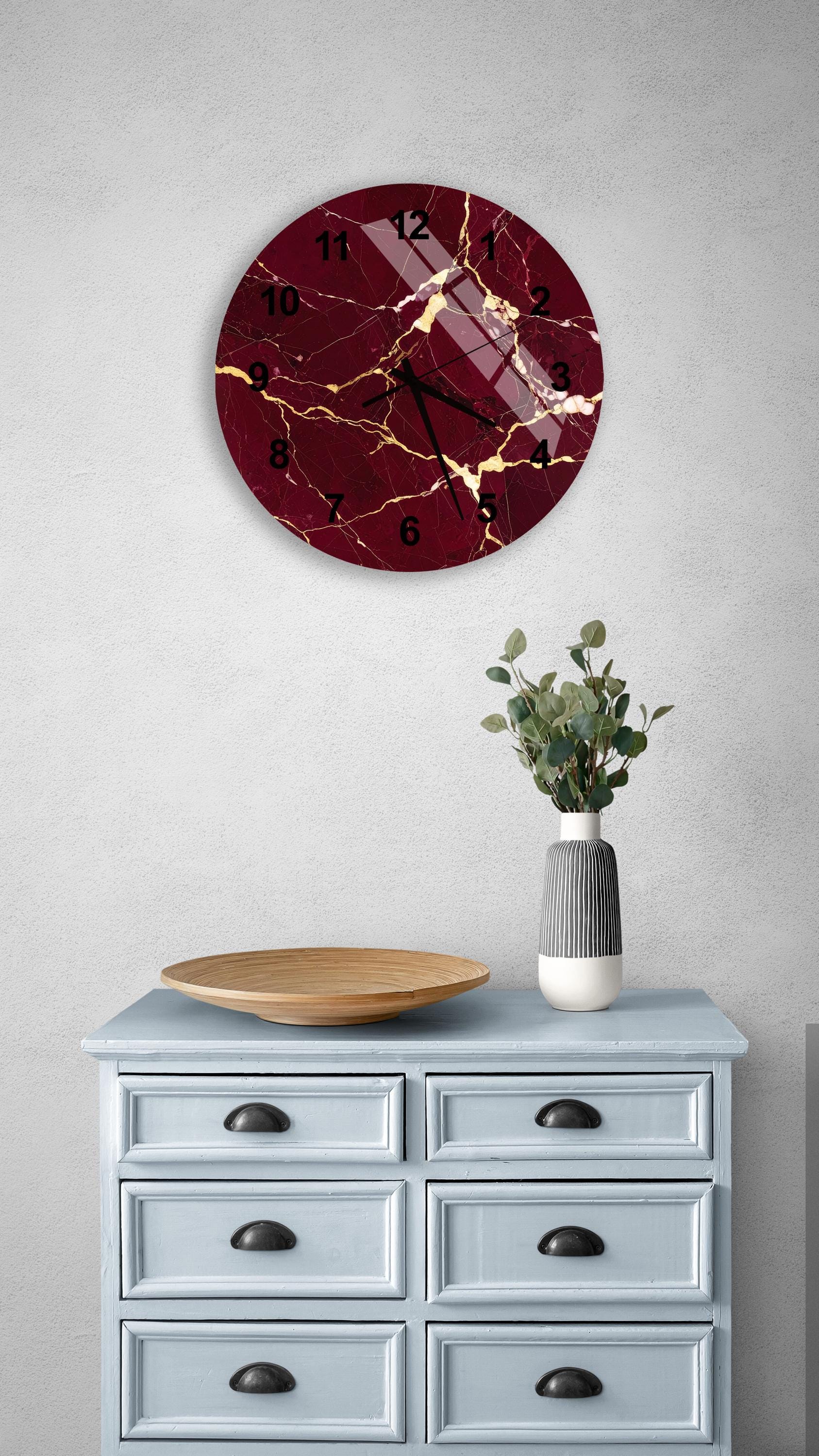 a wall clock with a maroon and gold design, mounted on a white wall above a light blue dresser with six drawers. The dresser also has a wooden bowl and a vase with greenery on it.