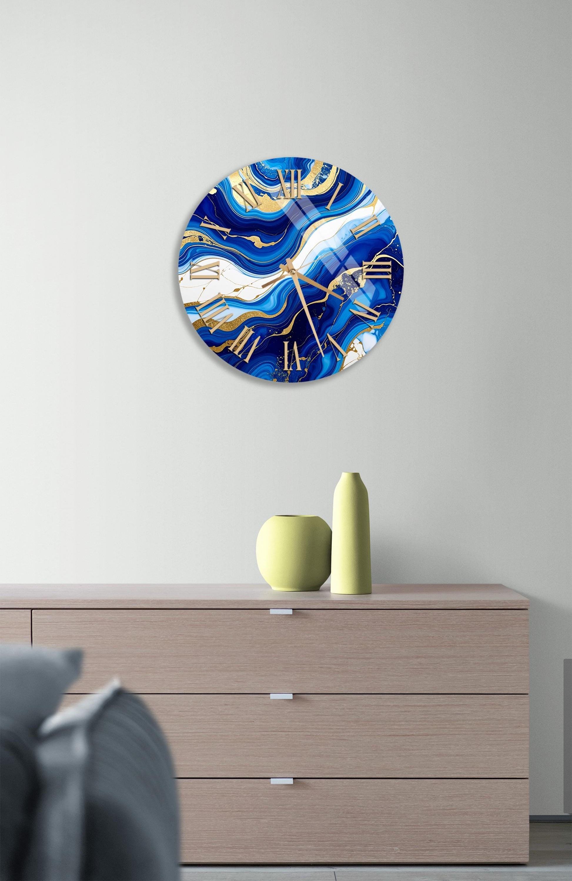 a modern clock with a blue and gold design, mounted on a wall above a wooden dresser. The dresser has a vase and a small sculpture on top of it.
