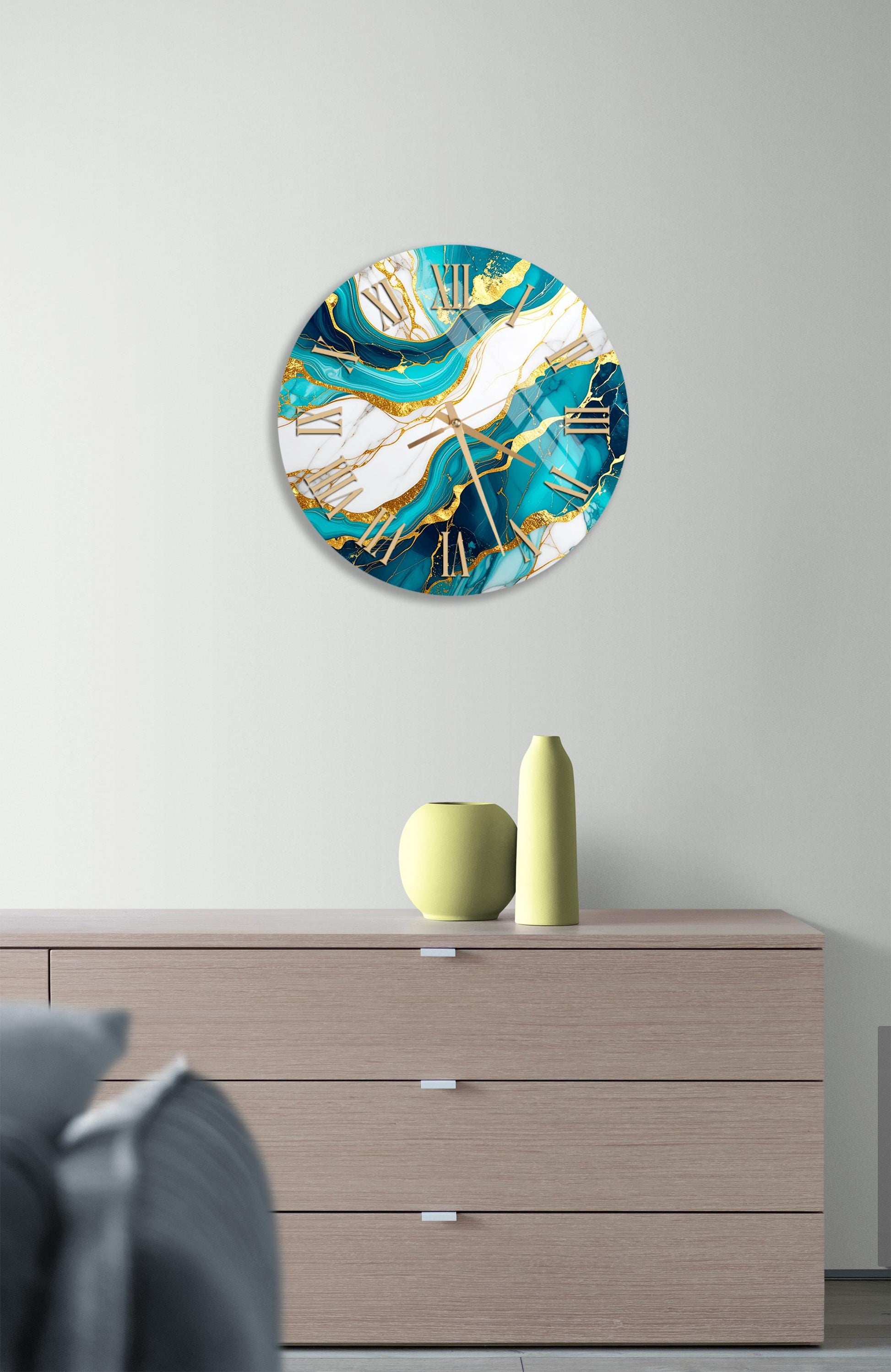 a modern clock with a turquoise and white color scheme, hanging on a wall above a wooden dresser. The dresser has a vase and a small sculpture on top of it.