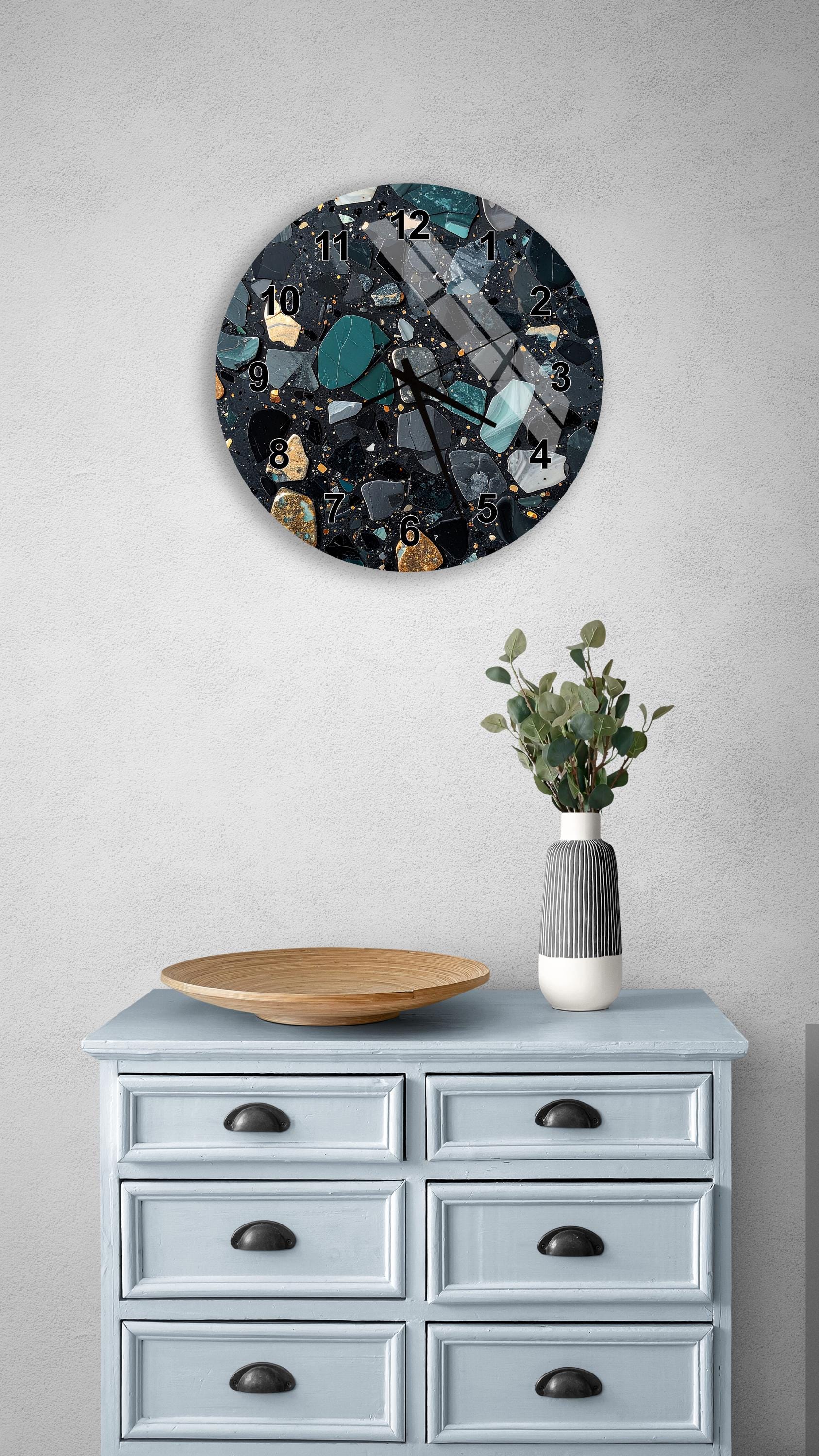 a wall clock with a black and white design, mounted on a wall above a light blue dresser with six drawers. The dresser also has a wooden bowl and a vase with greenery on it.