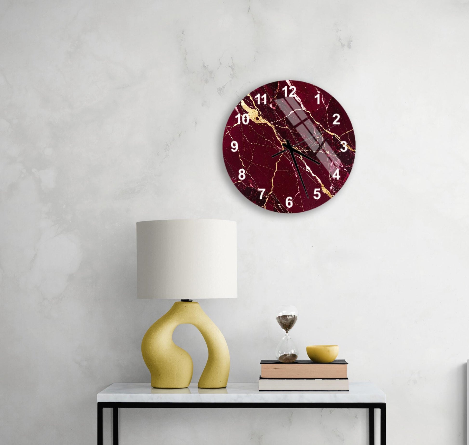 a white marble wall with a round clock featuring a maroon and gold design, a yellow lamp with a unique curved base, and a small table with a book and a glass of wine.