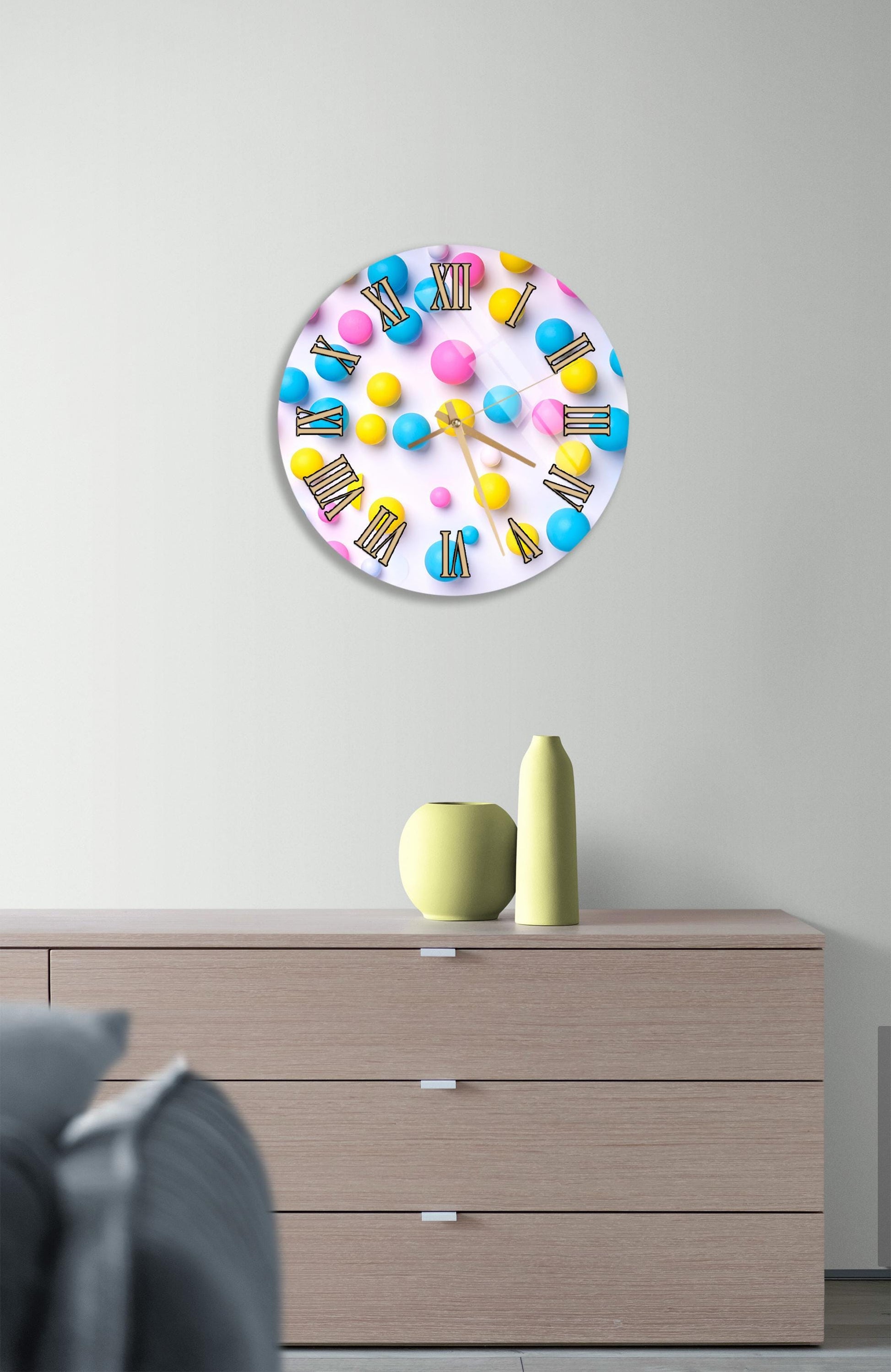 a round clock with colorful balls on a white background, mounted on a wall above a wooden dresser with three drawers.