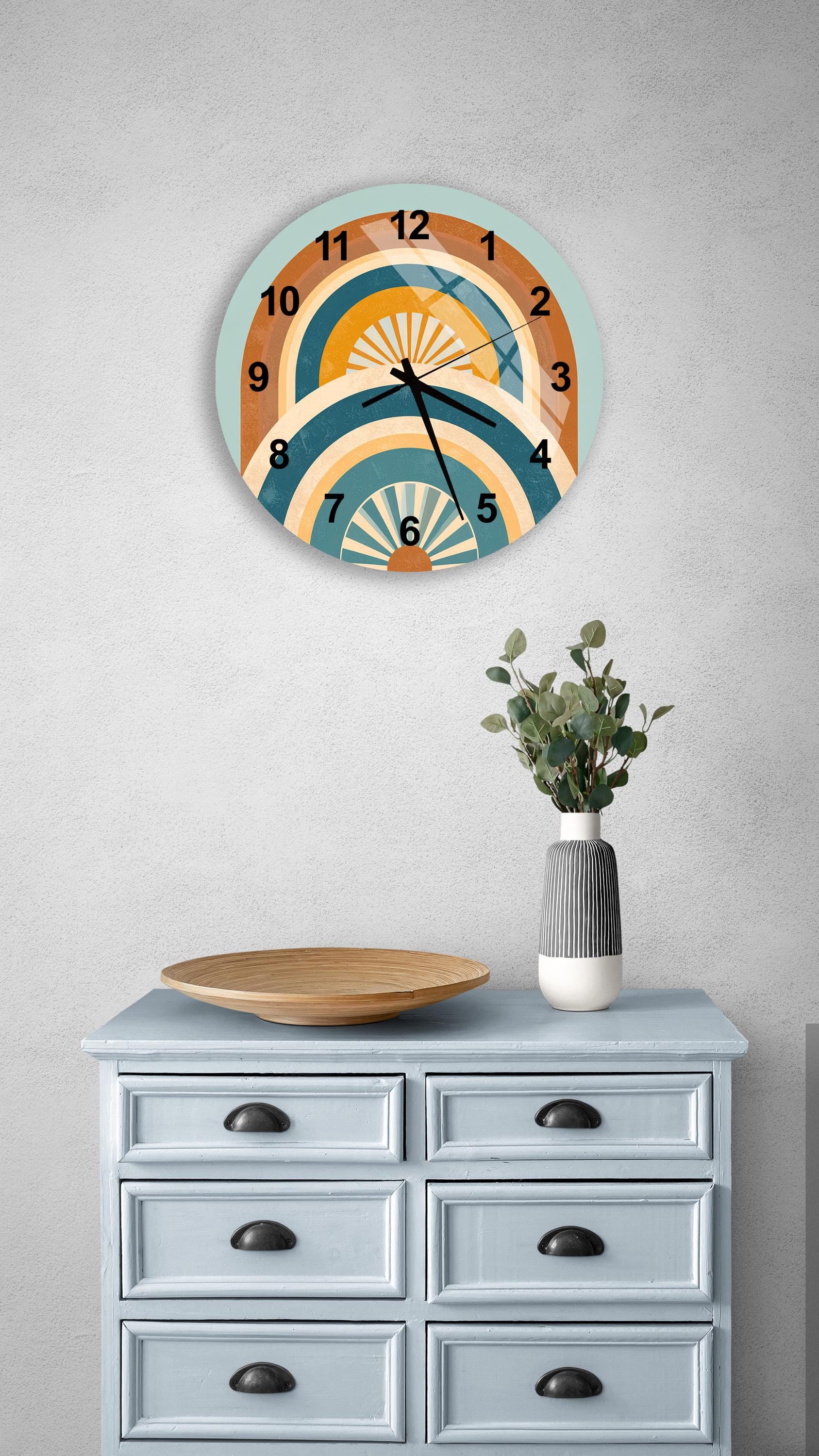 a wall clock with a colorful design, a wooden tray, a vase with greenery, and a gray dresser with six drawers.