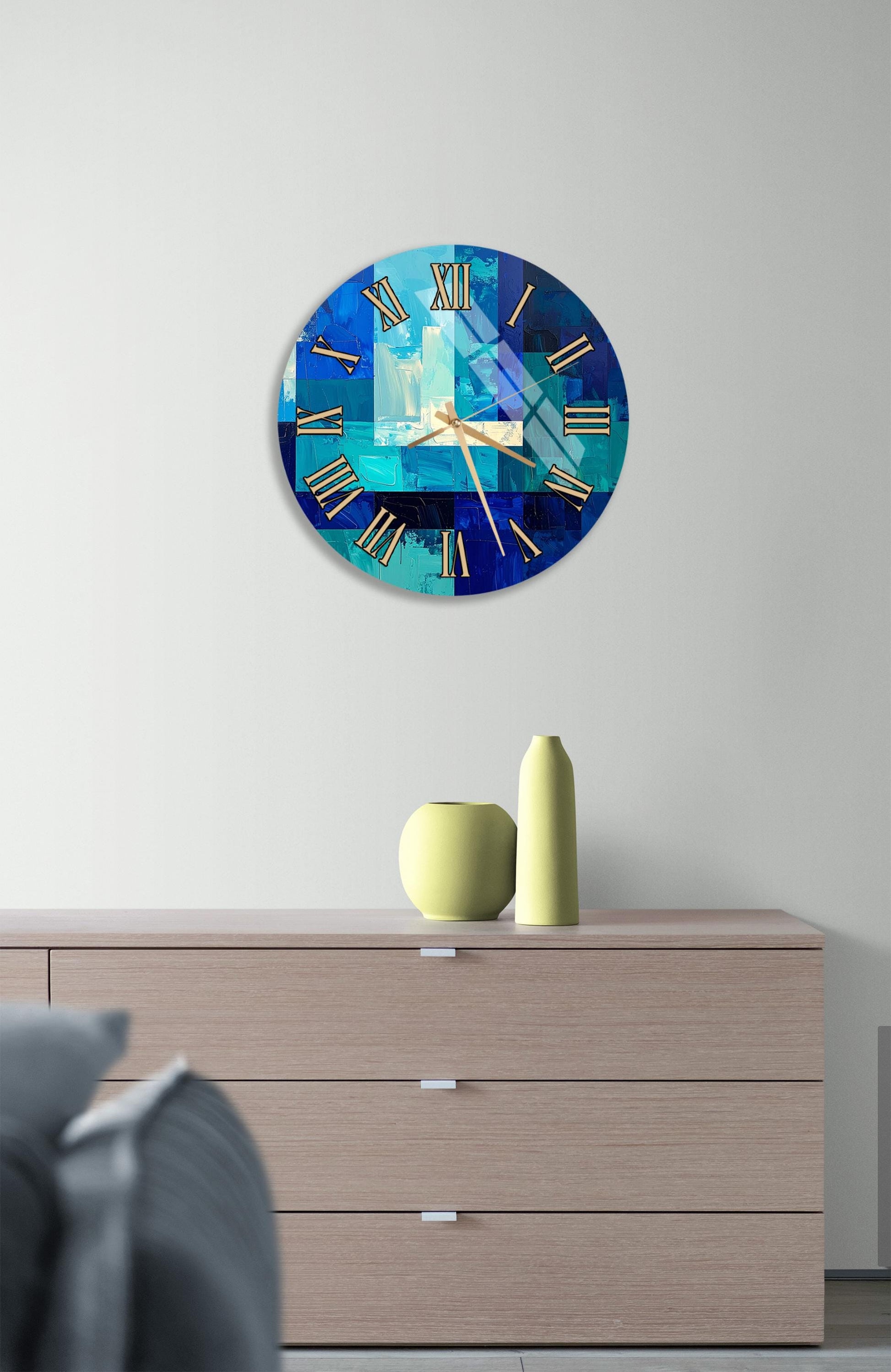 a modern clock with a blue and white color scheme, mounted on a wall above a wooden dresser. The dresser has a vase and a small sculpture on top of it, adding a decorative touch to the room.
