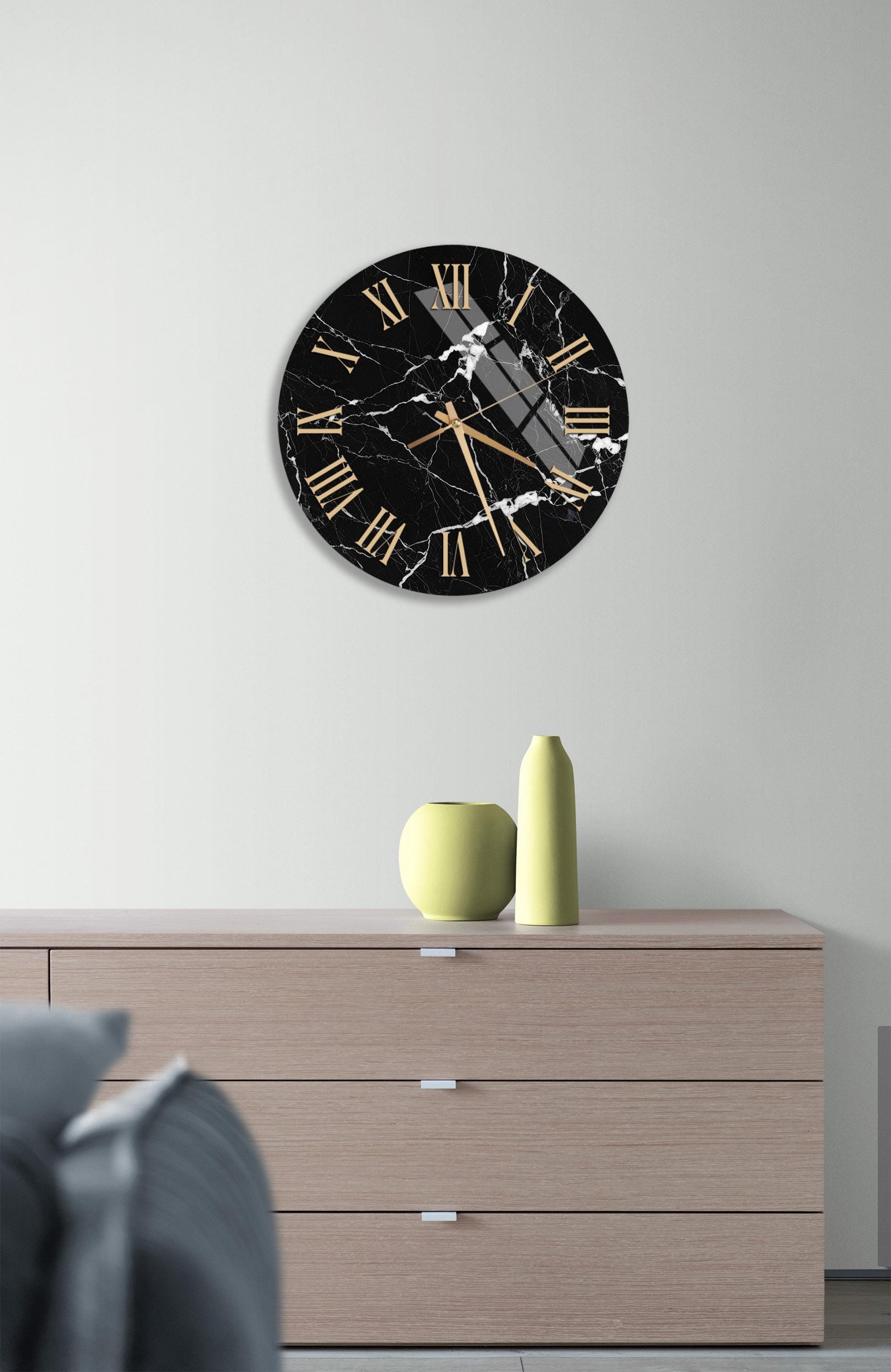 a black and white marble clock with gold Roman numerals, mounted on a white wall above a wooden dresser with three drawers.