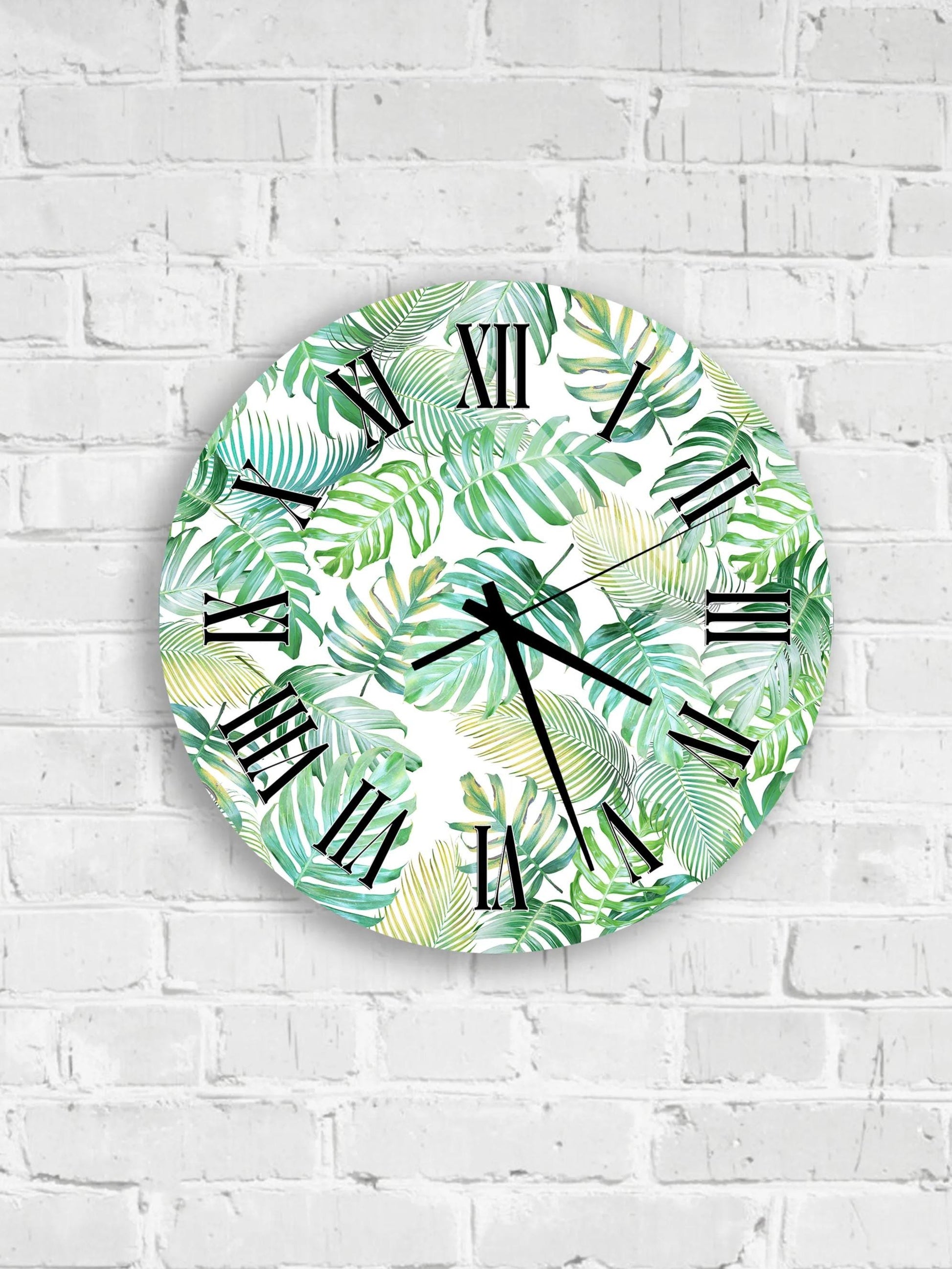 A round wall clock with a tropical leaf pattern on its face is mounted on a white brick wall.