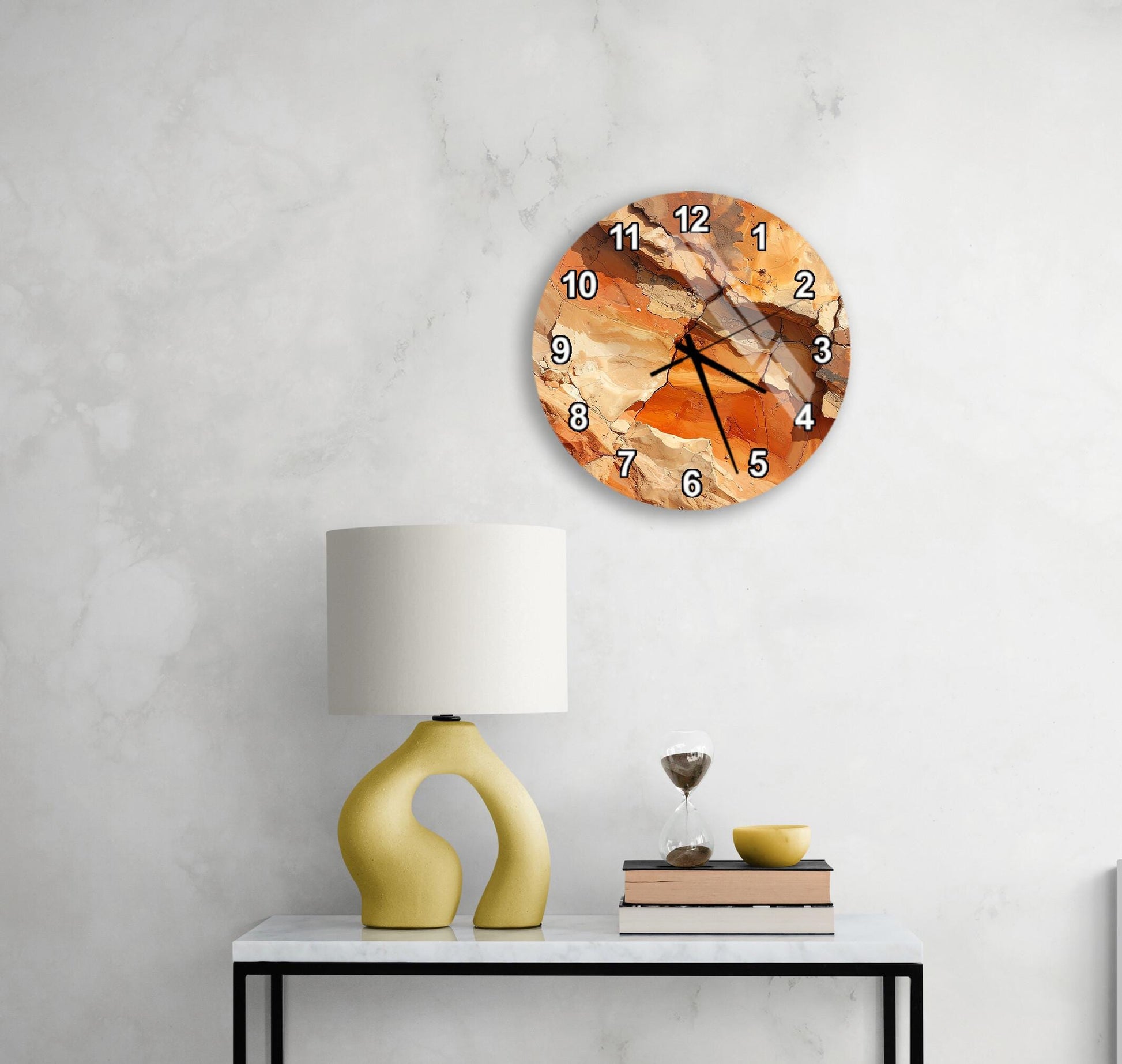 a modern wall clock with a unique orange and brown design, mounted on a white wall above a white table with a yellow lamp and a stack of books.