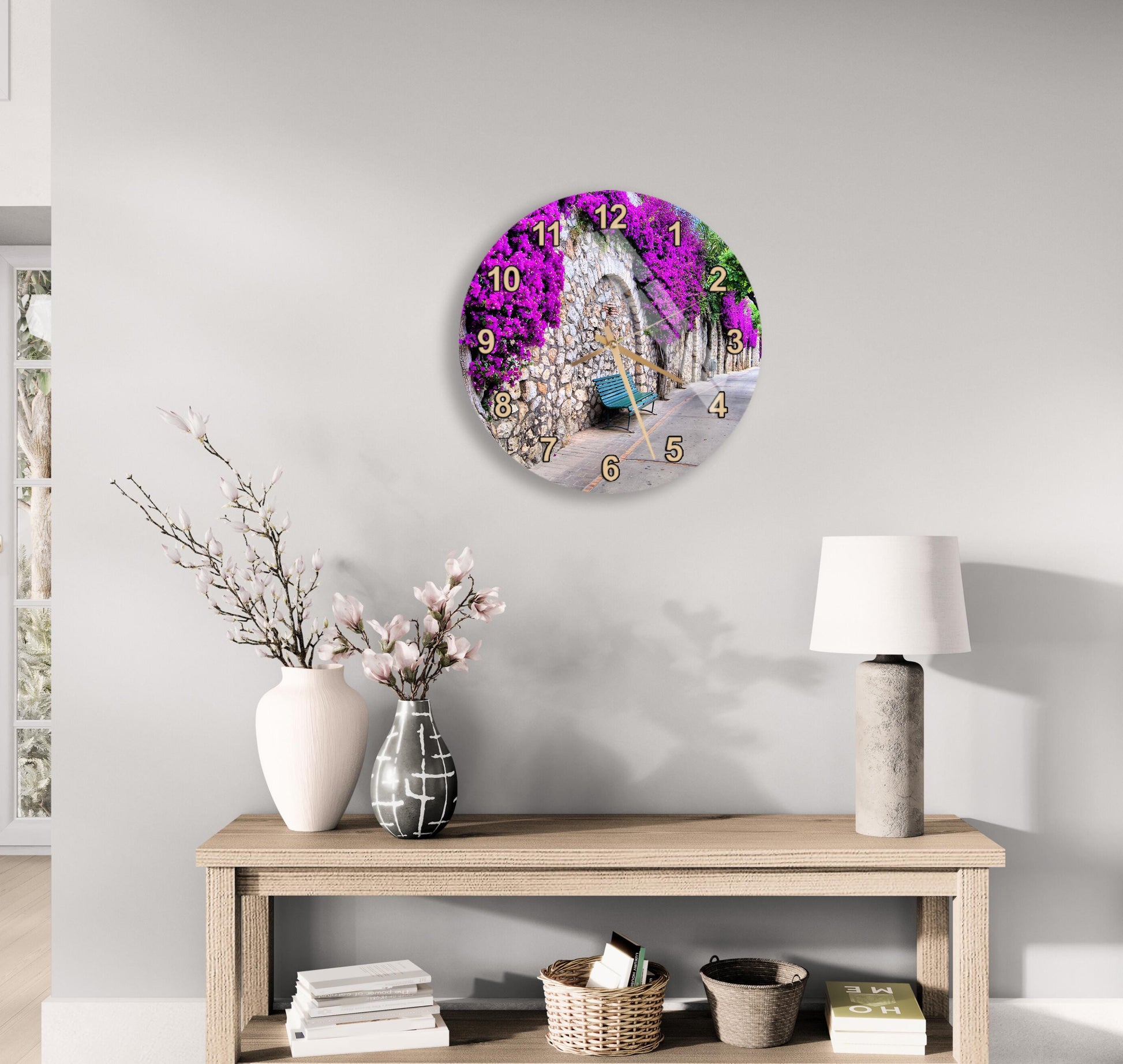 a wall clock with a purple floral design, a wooden table with a lamp, a vase with pink flowers, and a stack of books.