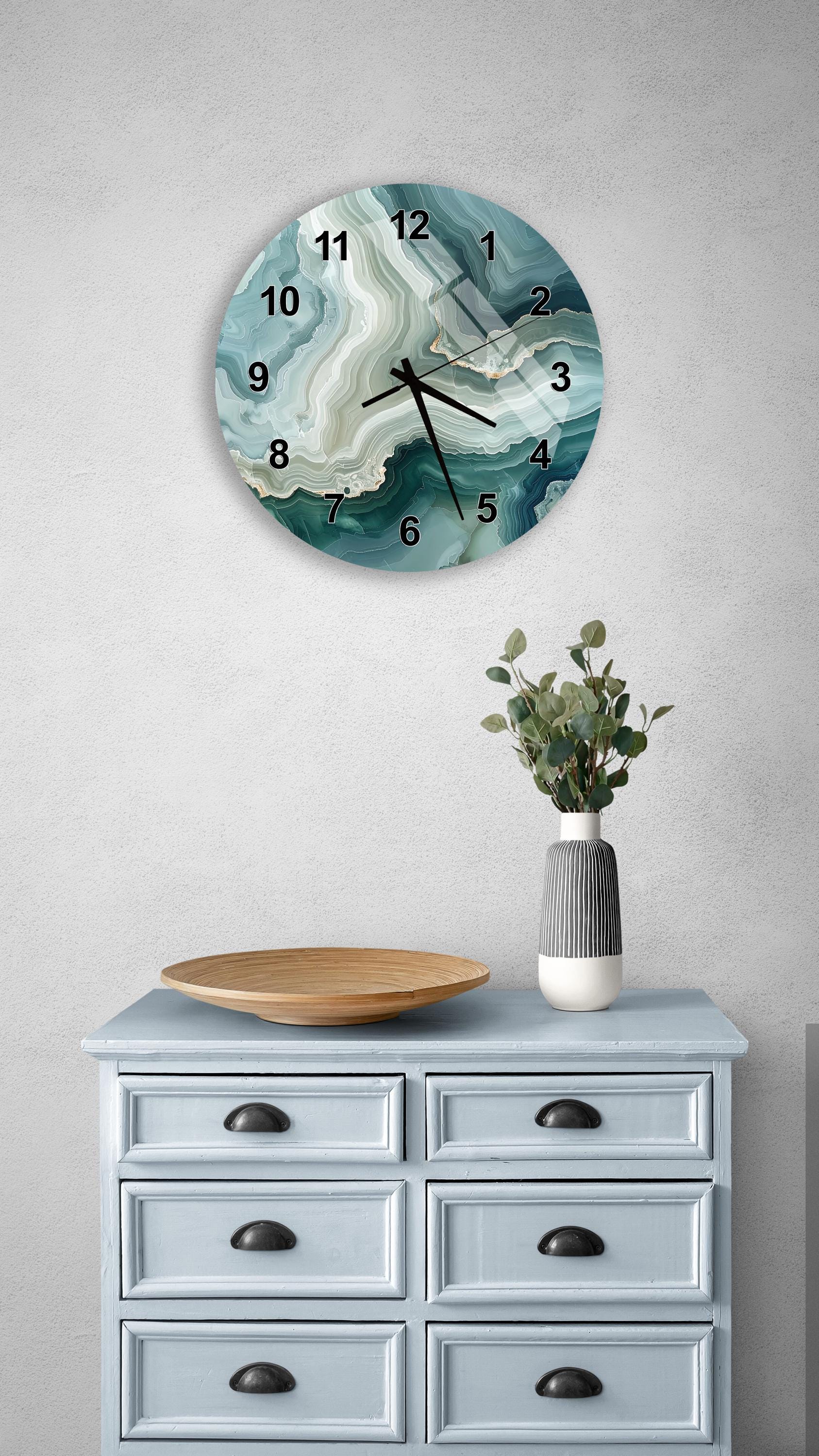 a wall clock with a marble-like design, mounted on a wall above a light blue wooden dresser with six drawers. The dresser also has a vase with greenery and a wooden tray on top.