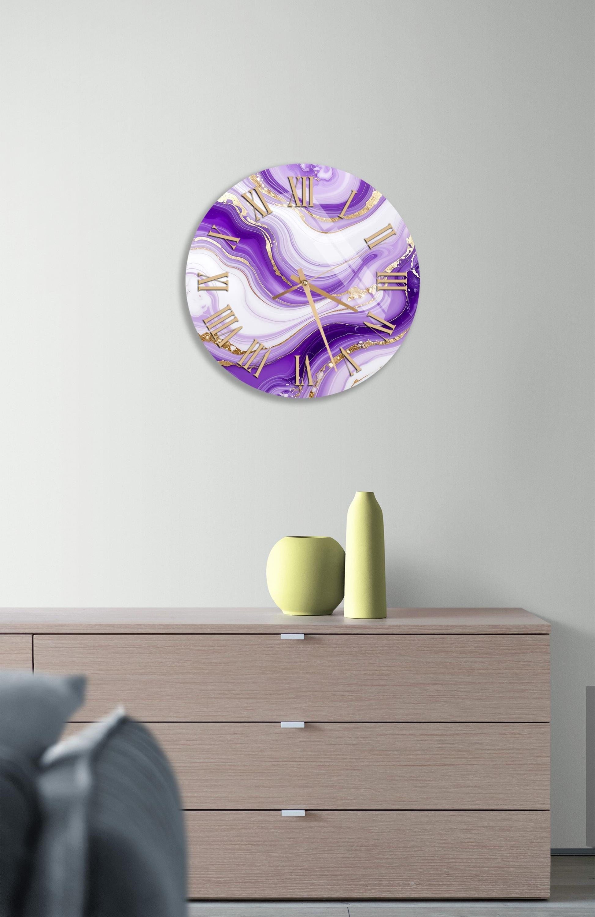 a modern interior design with a large round clock with a purple and white marble-like pattern on the wall above a wooden dresser. The dresser has three drawers and a small yellow vase on top.