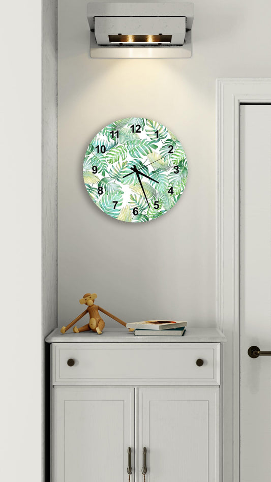 a wall clock with a tropical leaf pattern, mounted on a white wall above a white cabinet.