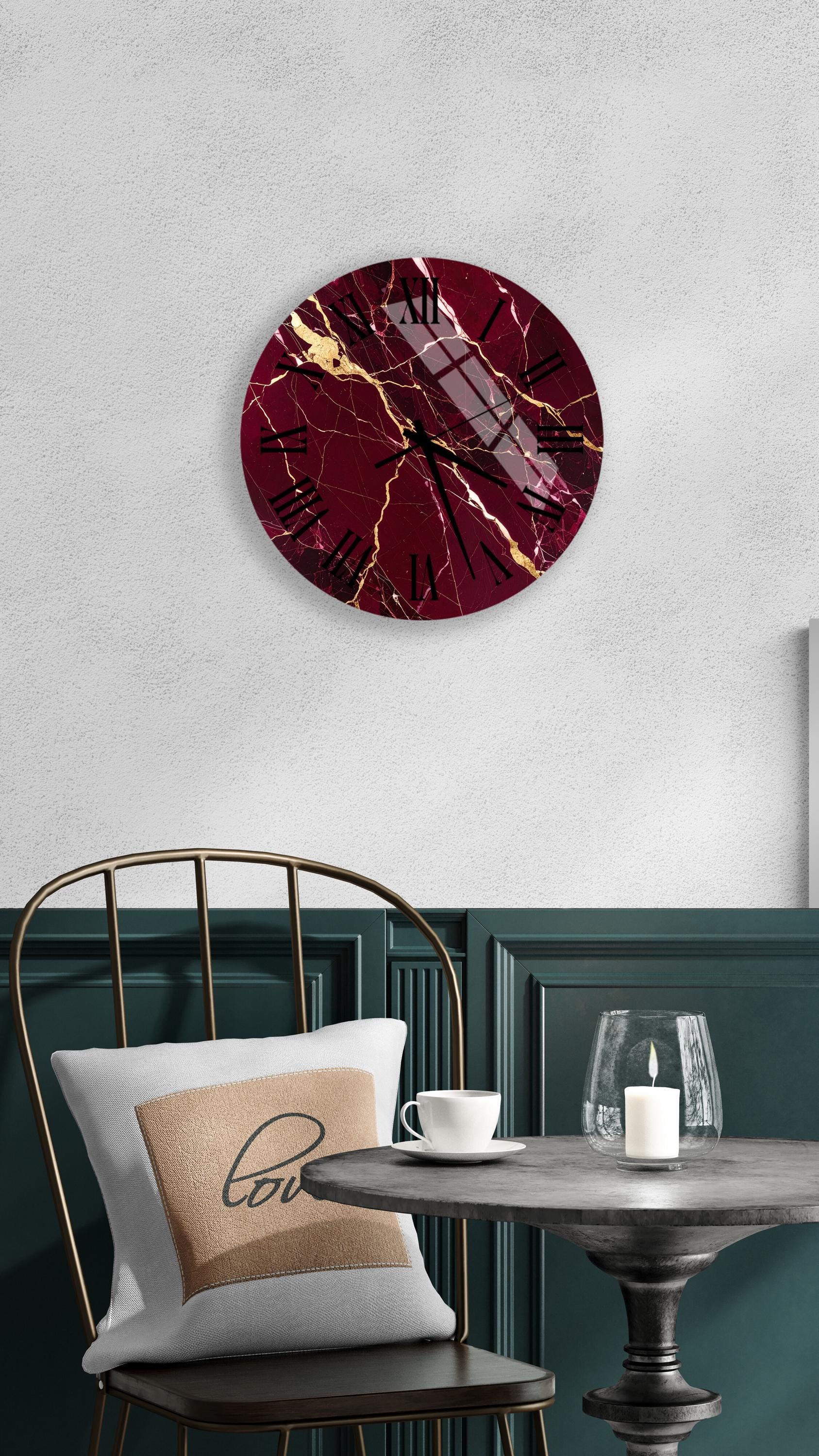 a modern interior design with a maroon and gold marble clock mounted on a white wall above a black metal chair. The chair has a white pillow with the word "love" printed on it. In front of the chair, there is a small table with a glass candle holder and a white teacup.