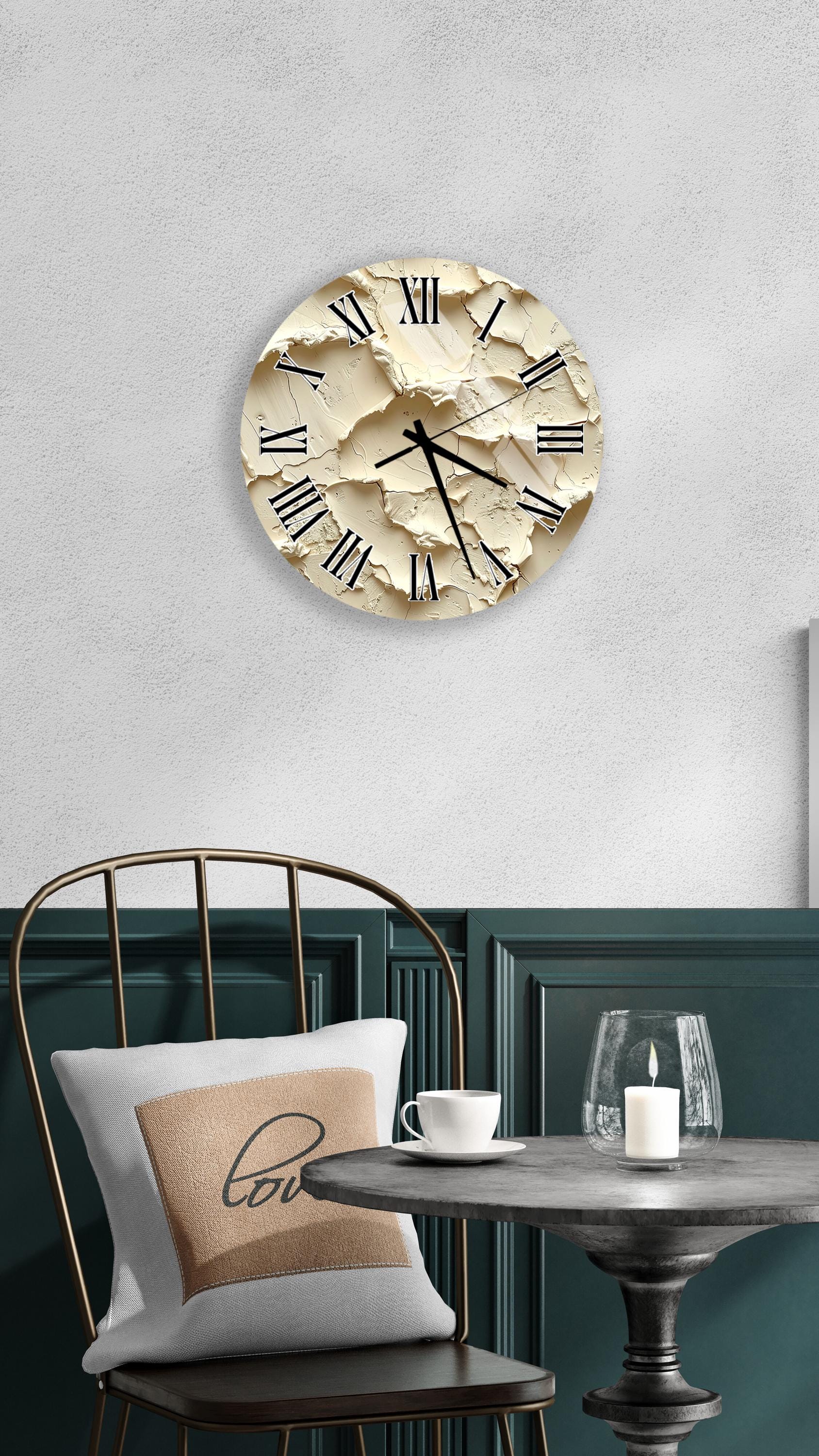 a round clock with a beige face and black Roman numerals mounted on a white wall above a black metal chair with a white pillow. The chair is positioned in front of a small table with a glass and a candle on it.