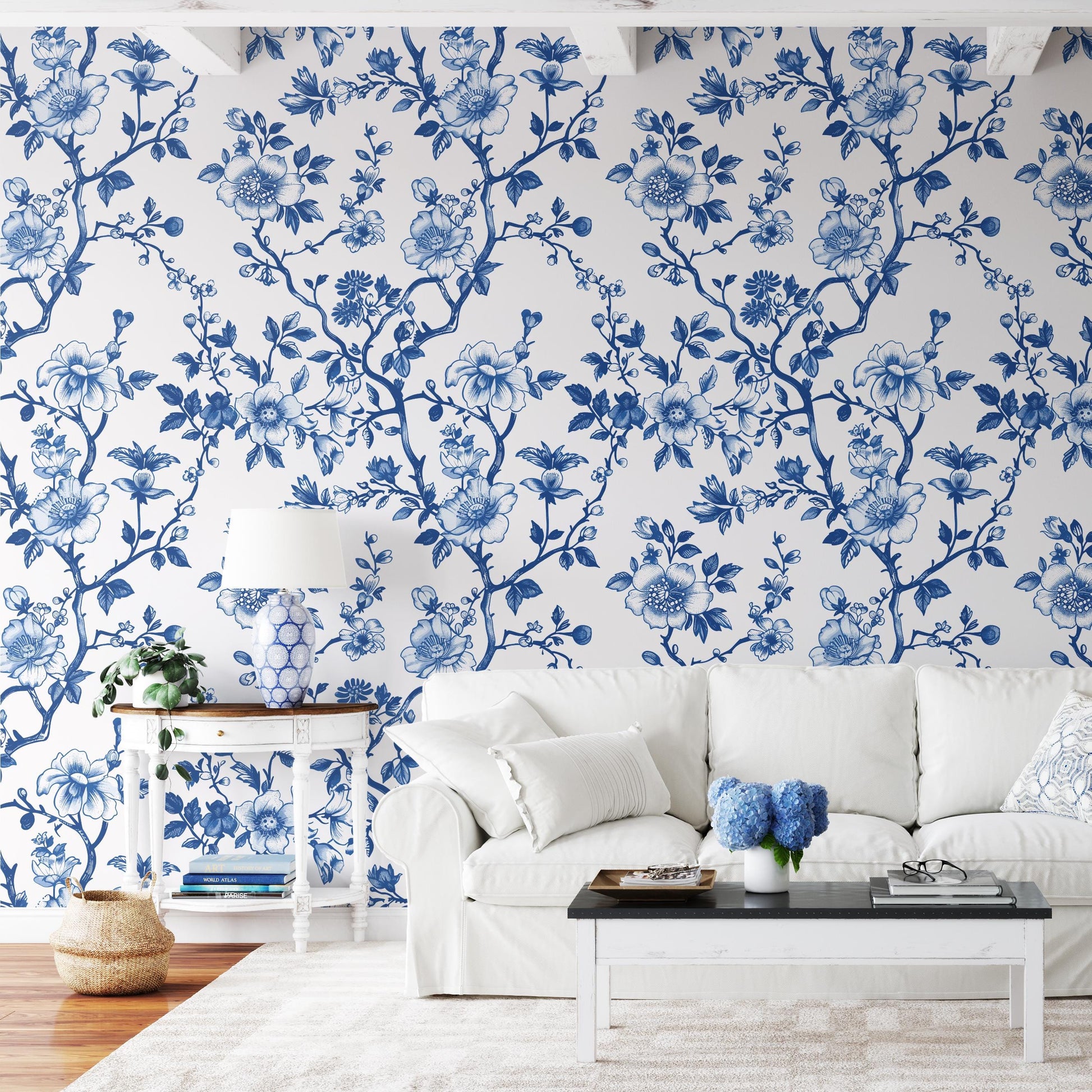 a living room with a white sofa, a coffee table, and a lamp. The walls are covered in a blue floral wallpaper, and there are some books and a vase on the coffee table.