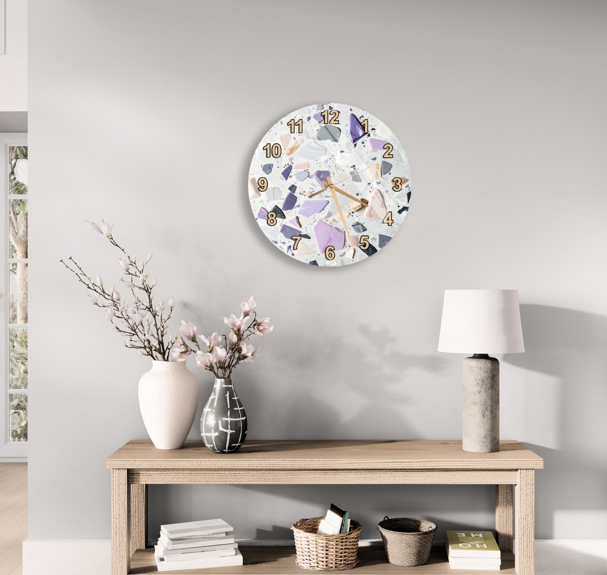 a modern and minimalist living room with a round wall clock displaying the time as 12:30. The clock is mounted above a wooden table with a lamp, a vase with flowers, and a stack of books. The table also has a basket and a small plant. The room has a neutral color scheme with white walls and a gray floor, creating a calm and serene atmosphere.