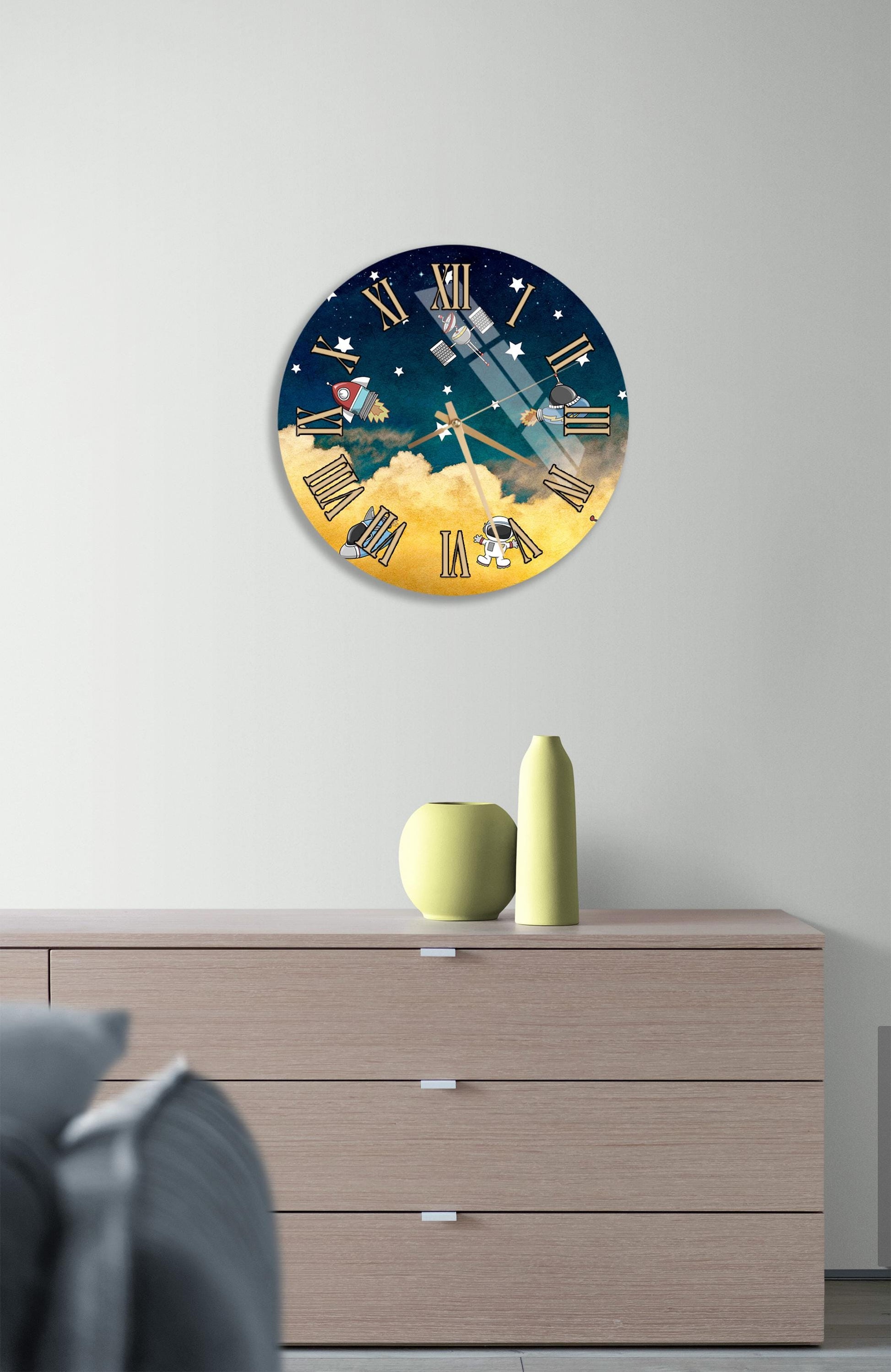 a wall clock with a space-themed design, mounted on a wall above a wooden dresser with a vase and a small sculpture on top.