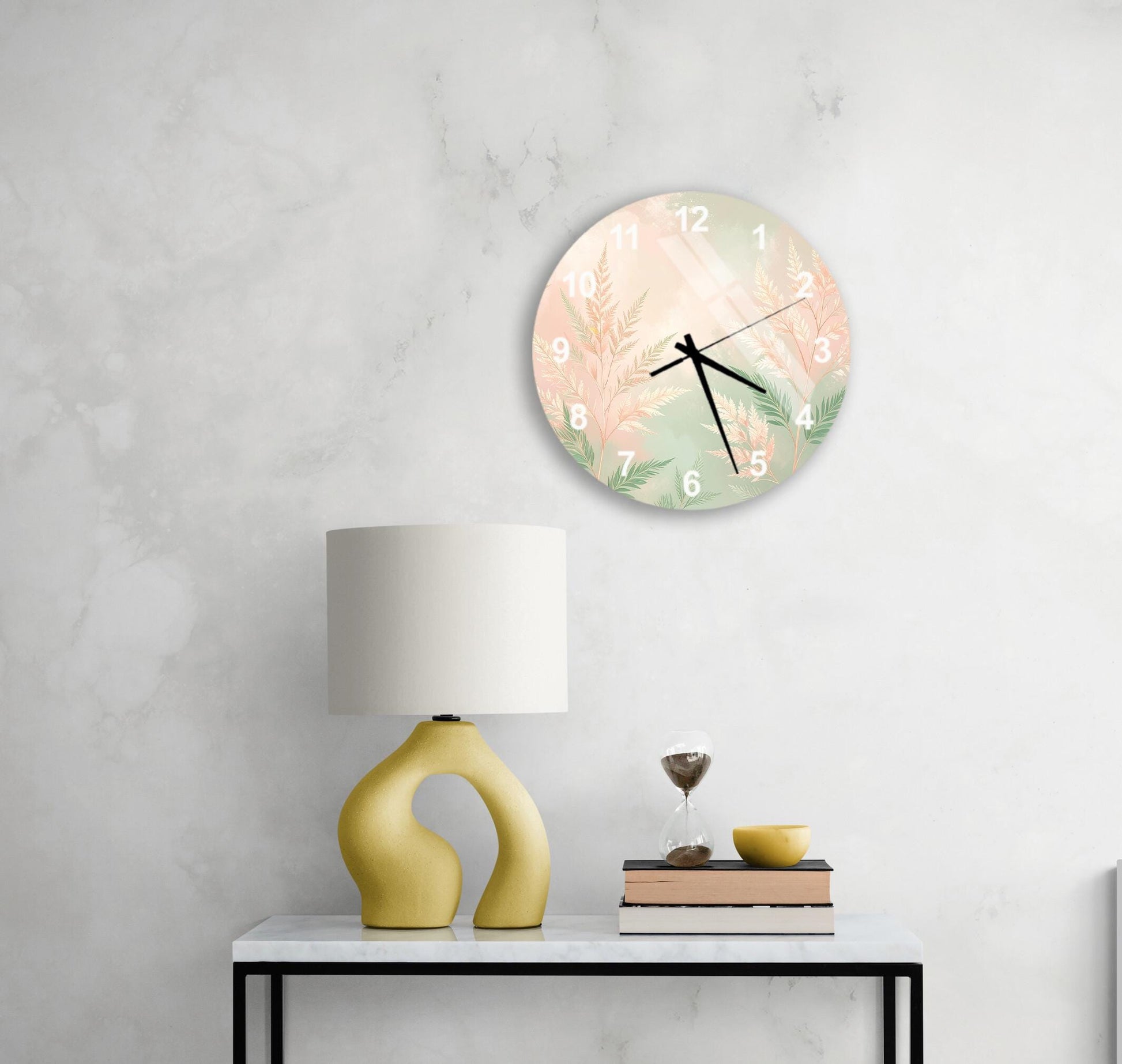 a white wall with a round clock displaying a floral design, a yellow lamp with a unique curved base, a stack of books, and a glass of wine on a white table.