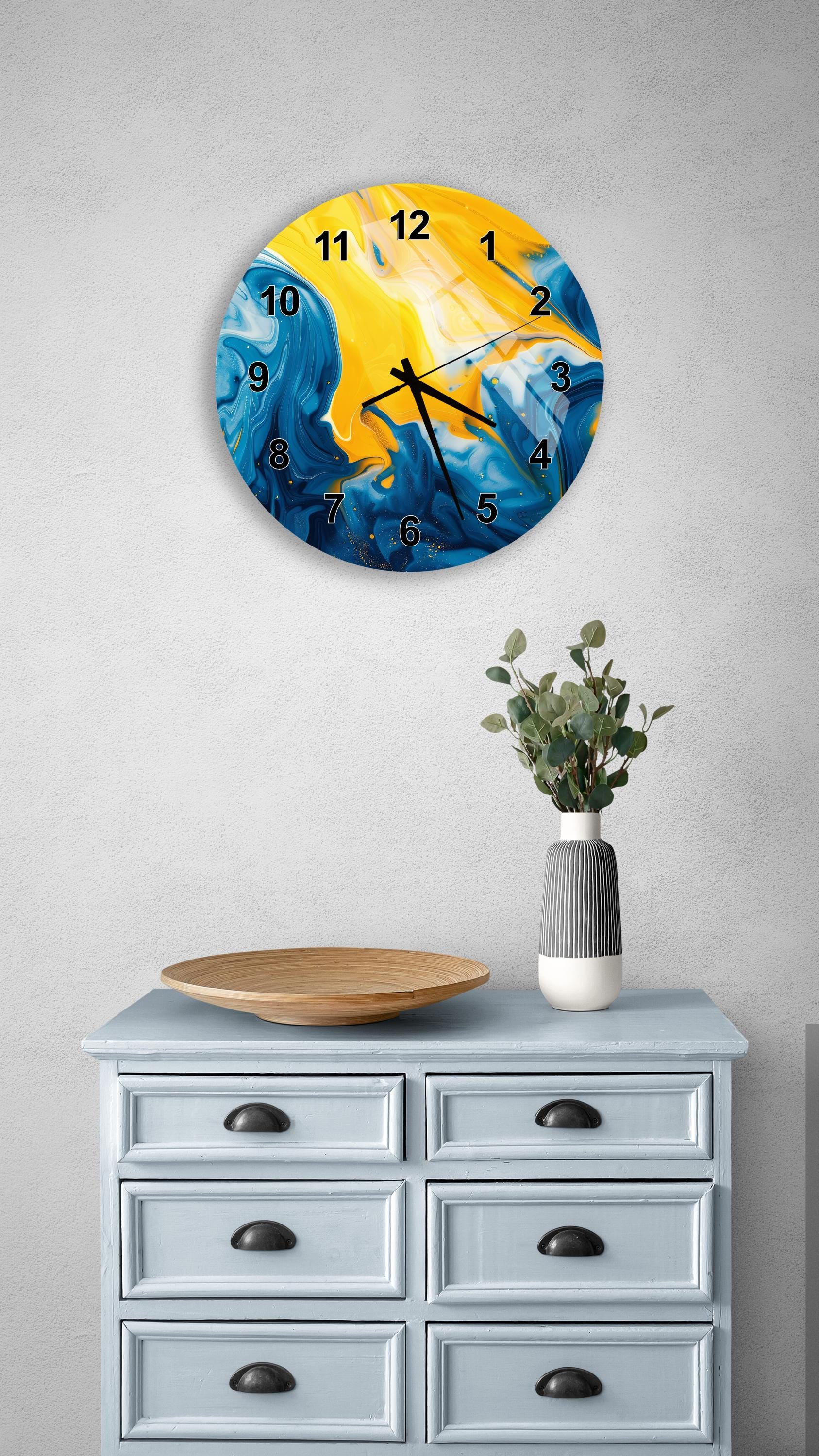 a wall clock with a vibrant blue and yellow design, mounted on a wall above a light blue dresser with six drawers. The dresser also has a vase with greenery on it, and there is a wooden plate on the dresser.